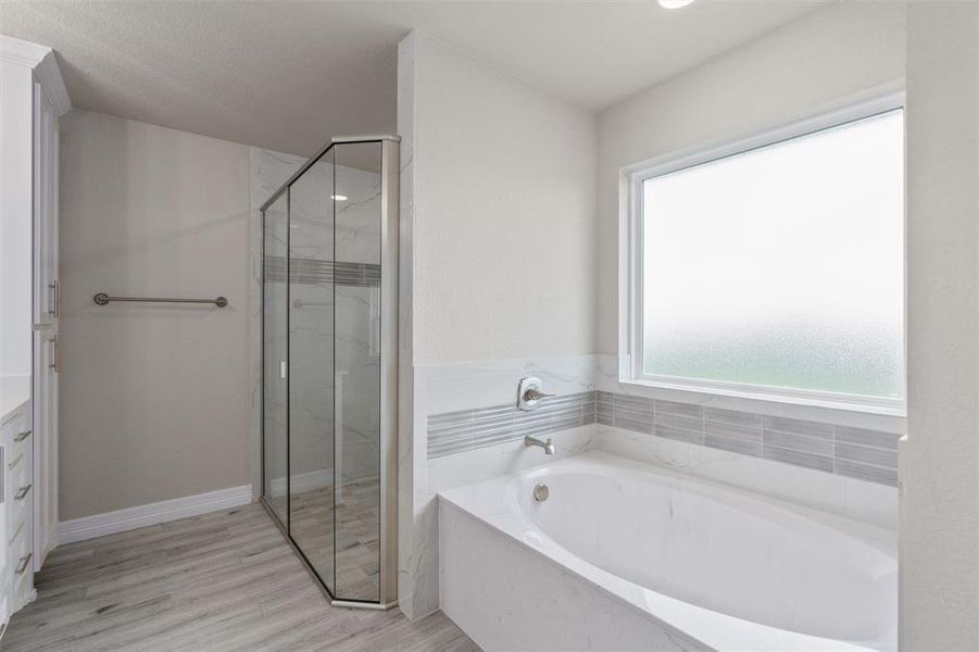 Soaking tub with a tiled surround, frosted window, glass-enclosed shower, wood-finish flooring, and white cabinetry Soaking tub with a tiled surround, frosted window, glass-enclosed shower, wood-finish flooring, and white cabinetry