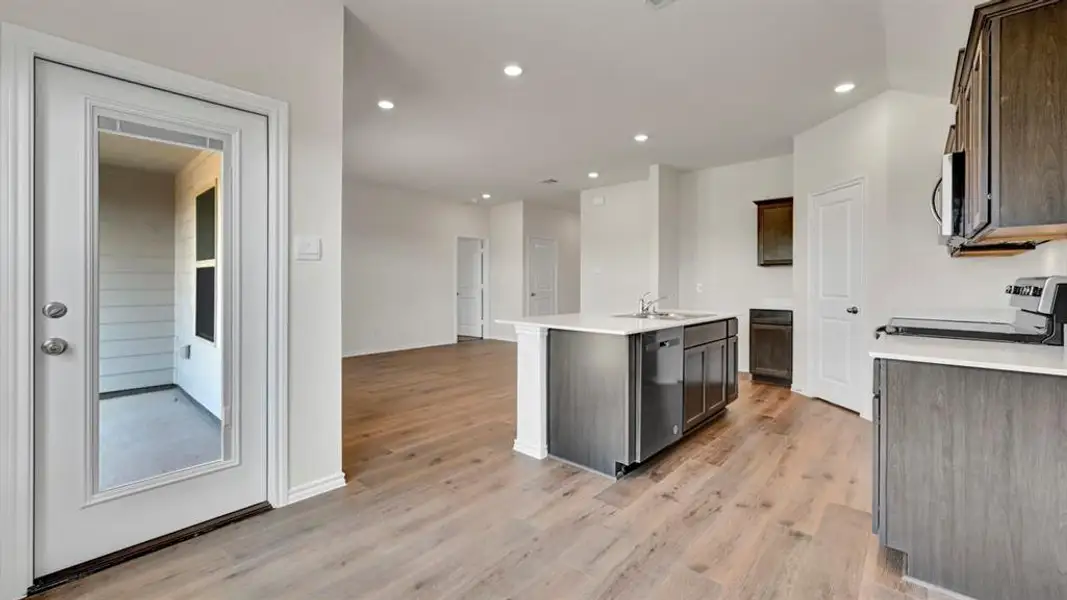 Open-concept kitchen and living area featuring wood-finish flooring throughout, recessed lighting, a kitchen island with an integrated sink and dishwasher, and dark wood cabinetry with light countertops