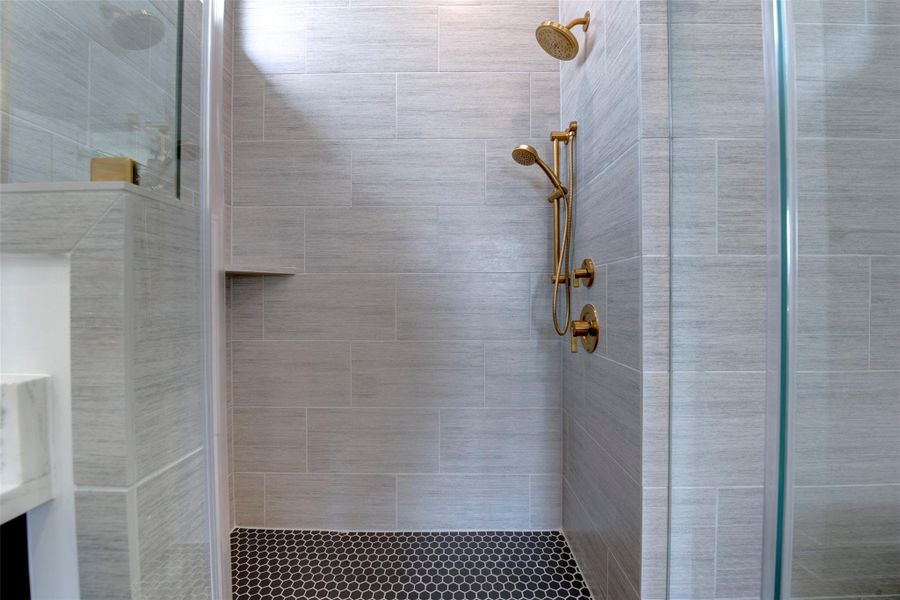 This photo showcases a modern bathroom featuring a spacious walk-in shower with elegant marble tiles and a stylish gold shower fixture. The space is complemented by a sleek vanity with a marble countertop and contemporary lighting.