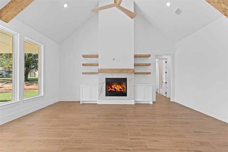 Unfurnished living room featuring light wood-type flooring, high vaulted ceiling, a premium fireplace, beam ceiling, and a ceiling fan