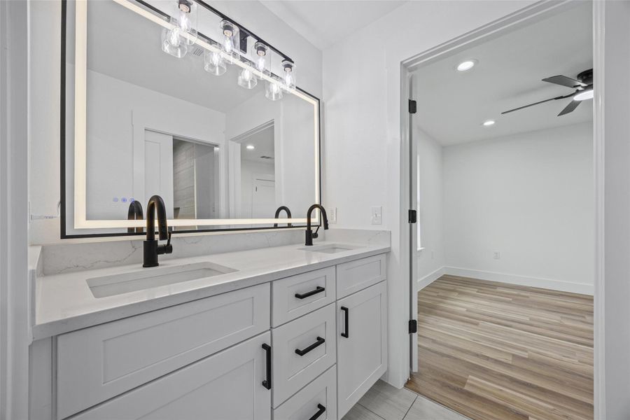 Jack and Jill bath. Dual vanity with light wood-finish flooring, quartz-finish countertops, matte black hardware, integrated backlit mirror, and a modern light fixture Jack and Jill bath. Dual vanity with light wood-finish flooring, quartz-finish countertops, matte black hardware, integrated backlit mirror, and a modern light fixture