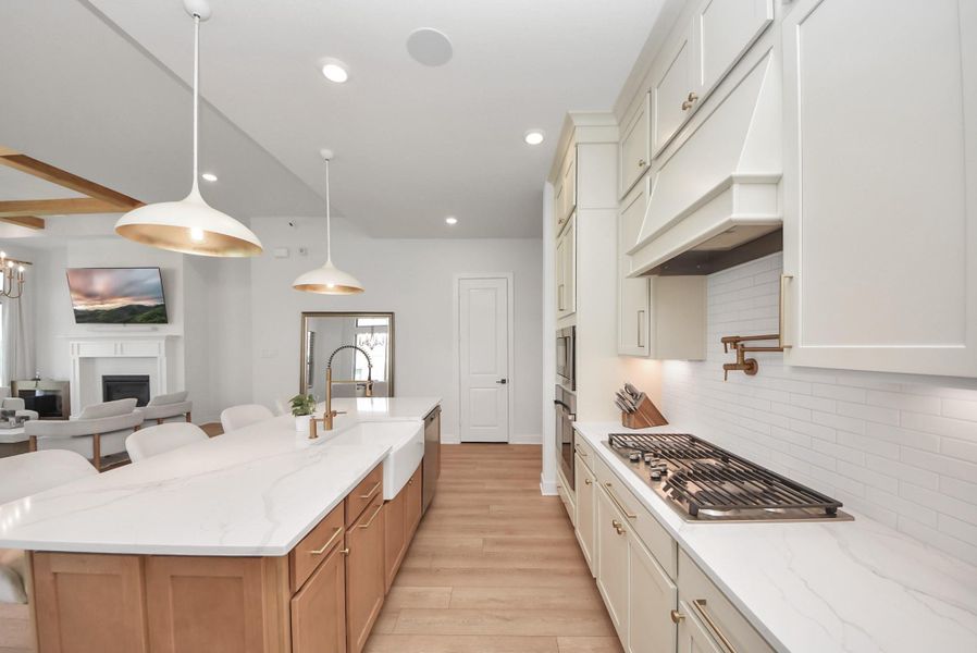Modern kitchen with sleek white cabinetry, elegant pendant lighting, and a spacious island with seating. A cozy living area with a fireplace and TV is visible, creating a perfect open-concept space for entertaining.