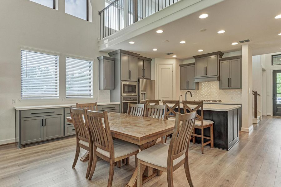 This photo showcases a spacious, open-concept kitchen and dining area with high ceilings. The kitchen features modern gray cabinetry, stainless steel appliances, and a large island with seating. Ample natural light streams through two large windows, highlighting the elegant wood flooring and contemporary design.