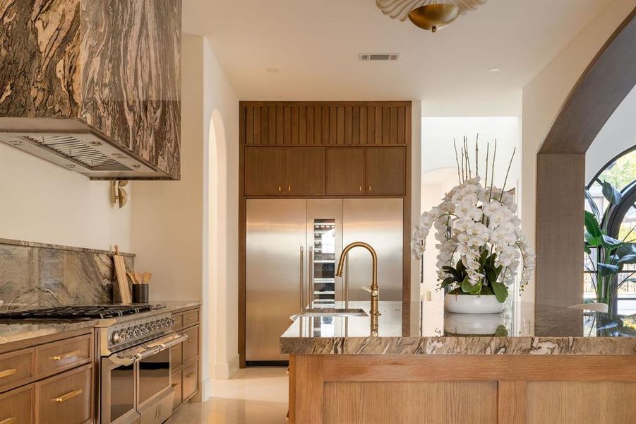 Kitchen with high quality appliances, custom range hood, light stone counters, light tile patterned floors, and brown cabinetry