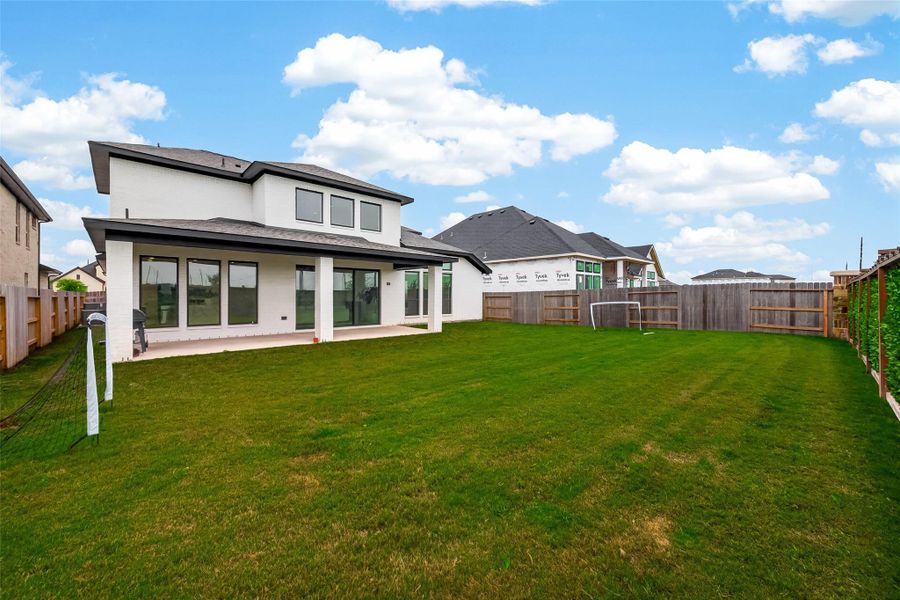 This photo showcases a spacious backyard with a well-maintained lawn, perfect for outdoor activities. It features a modern two-story home with large windows and a covered patio, ideal for relaxation and entertaining. The yard is enclosed with a wooden fence, offering privacy and security.