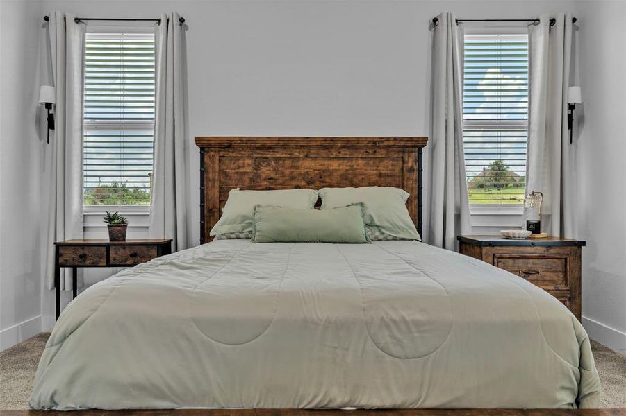 Bedroom featuring a wood-finish headboard, twin windows with blinds and drapery, and wall-mounted sconces