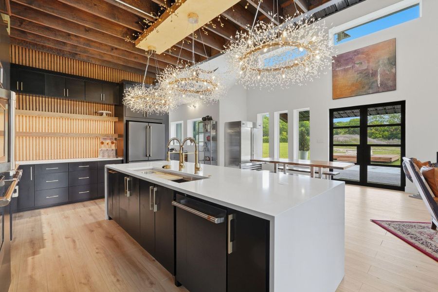 This modern kitchen features elegant black cabinetry, a spacious white island with dual sinks, and stunning decorative lighting. High ceilings with exposed beams, large windows, and glass doors provide ample natural light and views of the greenery outside. Perfect for entertaining, it seamlessly connects to a dining area with a sleek, open design.