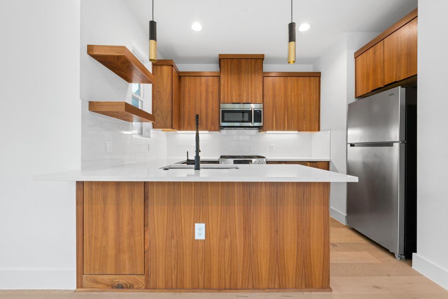 Kitchen with stainless steel appliances, open shelves, brown cabinetry, a peninsula, and light countertops Kitchen with stainless steel appliances, open shelves, brown cabinetry, a peninsula, and light countertops