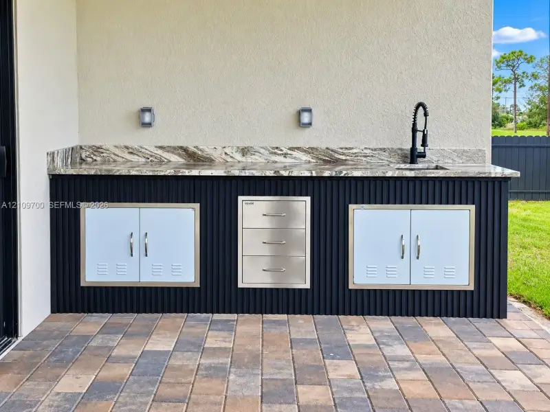 Outdoor kitchen featuring a stone-look countertop, integrated stainless steel storage drawers and cabinets, a high-arc pull-down faucet, and a fluted black exterior finish on a paver patio