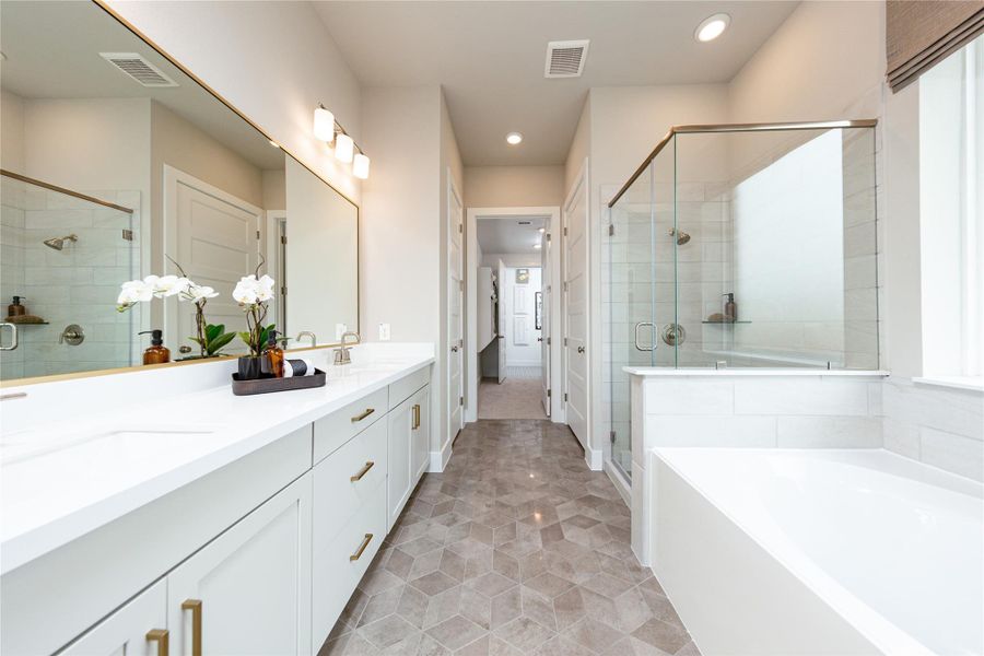 Luxurious primary suite bathroom featuring a spacious double vanity with crisp white countertops, modern gold-finished hardware, a frameless glass walk-in shower, and a relaxing soaking tub accented by elegant geometric tile flooring.