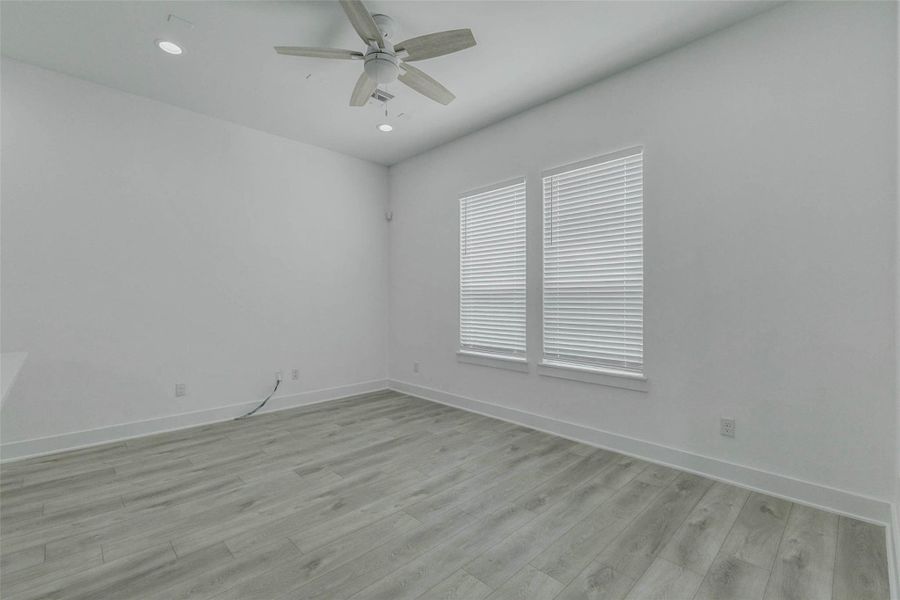 Spacious living room view with two large windows, white blinds, tall ceilings, ceiling fan, recessed lighting, and light wood-look floors. The neutral palette keeps the space fresh and inviting, while the open layout provides excellent flexibility for furniture placement, dining, lounging, or entertaining.