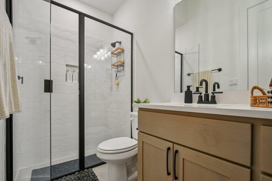 This bathroom features a shower with a black-framed glass enclosure and white geometric-patterned tile, complemented by a light wood vanity with a white countertop and black fixtures This bathroom features a shower with a black-framed glass enclosure and white geometric-patterned tile, complemented by a light wood vanity with a white countertop and black fixtures