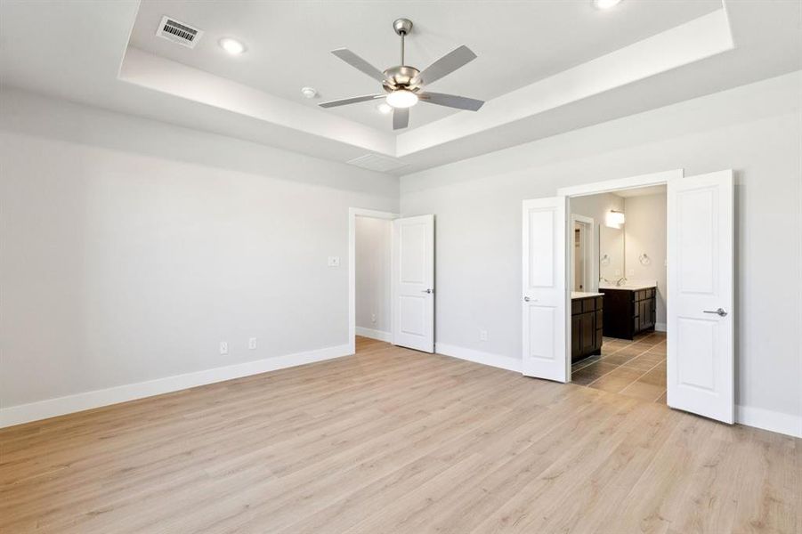 Unfurnished bedroom with a tray ceiling, light wood-style floors, a ceiling fan, ensuite bath, and recessed lighting Unfurnished bedroom with a tray ceiling, light wood-style floors, a ceiling fan, ensuite bath, and recessed lighting