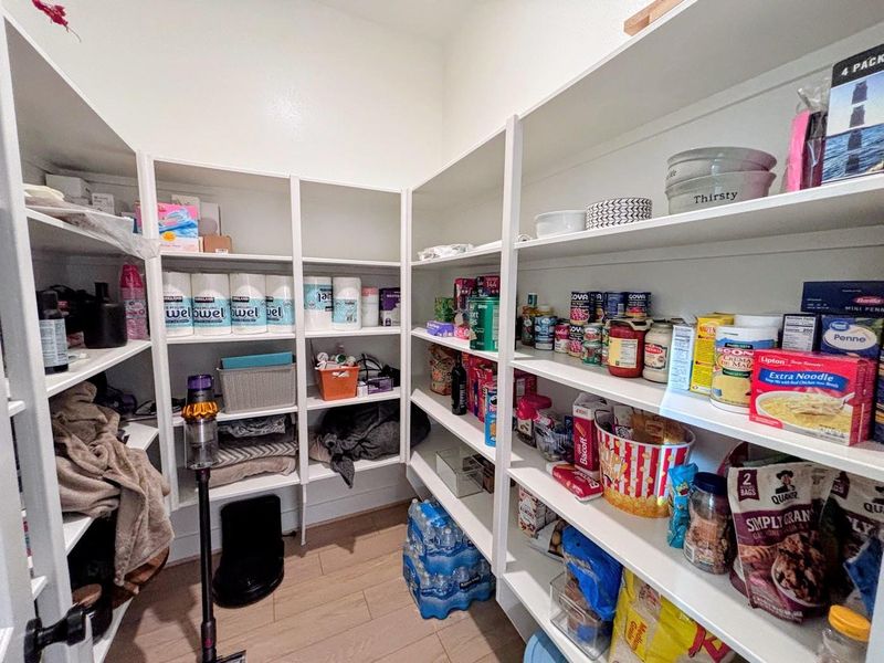 Spacious walk-in pantry with ample shelving on three walls, providing plenty of storage for pantry items, cleaning supplies, and household goods. The room is well-organized and brightly lit, with wood flooring enhancing its functionality and appeal.