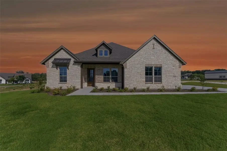 Stone facade residence featuring a dark shingle roof, a prominent dormer window, a covered entry, and a window awning Stone facade residence featuring a dark shingle roof, a prominent dormer window, a covered entry, and a window awning