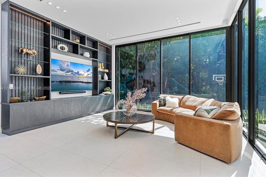 Living space featuring floor-to-ceiling glass walls, built-in custom dark wood cabinetry with integrated glass shelving, recessed lighting, and large-format tile flooring