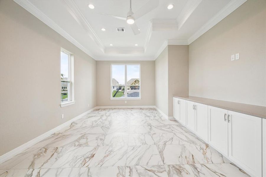 This is a spacious, well-lit room with elegant marble flooring, crisp white cabinetry, and a tray ceiling with recessed lighting. Large windows provide a view of the waterway.