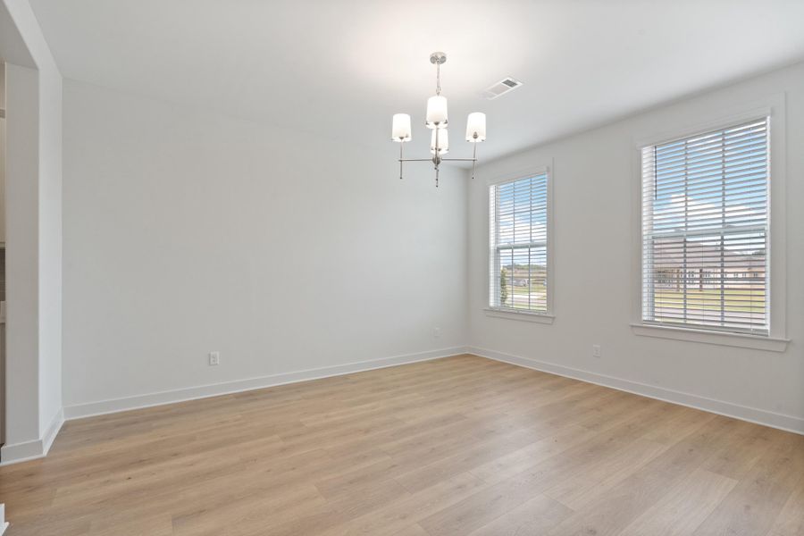 Spacious room featuring light wood-finish flooring, two large windows with blinds, a modern chandelier, and neutral wall tones Spacious room featuring light wood-finish flooring, two large windows with blinds, a modern chandelier, and neutral wall tones