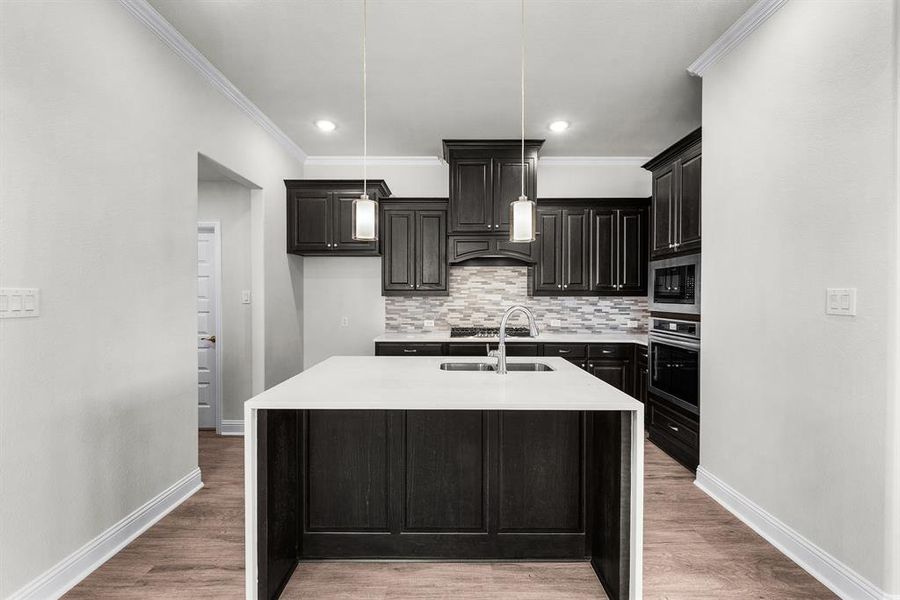 Kitchen with light countertops, ornamental molding, light wood-type flooring, stainless steel appliances, and a kitchen island with sink Kitchen with light countertops, ornamental molding, light wood-type flooring, stainless steel appliances, and a kitchen island with sink