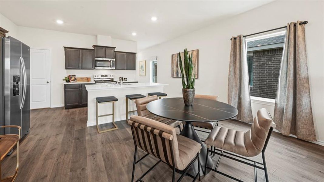 Open-concept kitchen and dining area featuring wood-finish flooring, dark wood cabinetry, stainless steel appliances, a white island with seating, and a window with a dark brick exterior beyond