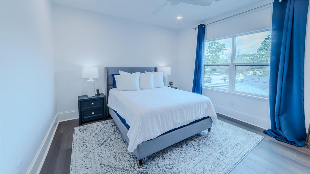 Bright and airy bedroom with large window, white walls, and blue accents. Features a cozy bed with a modern gray headboard, elegant nightstand, and a stylish area rug on wood flooring.