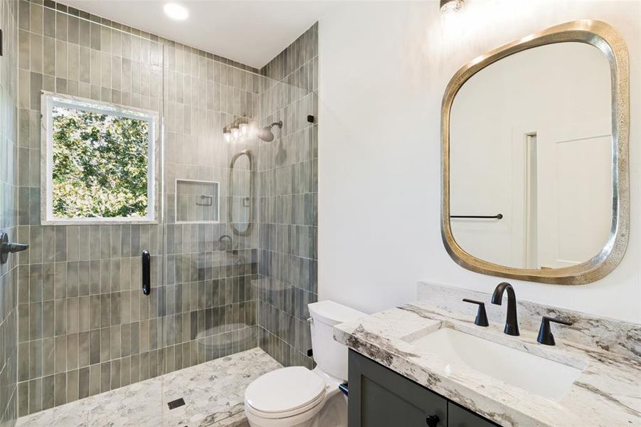 Bathroom featuring a walk-in shower with vertical tile and glass enclosure, a white vanity with stone countertop and integrated sink, a rectangular mirror, and matte black fixtures Bathroom featuring a walk-in shower with vertical tile and glass enclosure, a white vanity with stone countertop and integrated sink, a rectangular mirror, and matte black fixtures