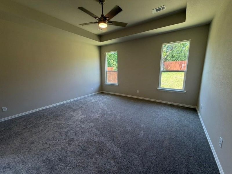 Spacious room featuring tray ceilings, a ceiling fan with integrated lighting, two windows, and neutral carpeting