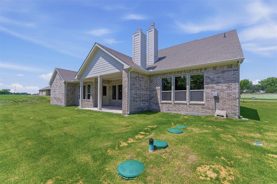 Rear exterior featuring a covered patio, extensive green lawn, brick and siding facade, multiple windows, and dual chimneys