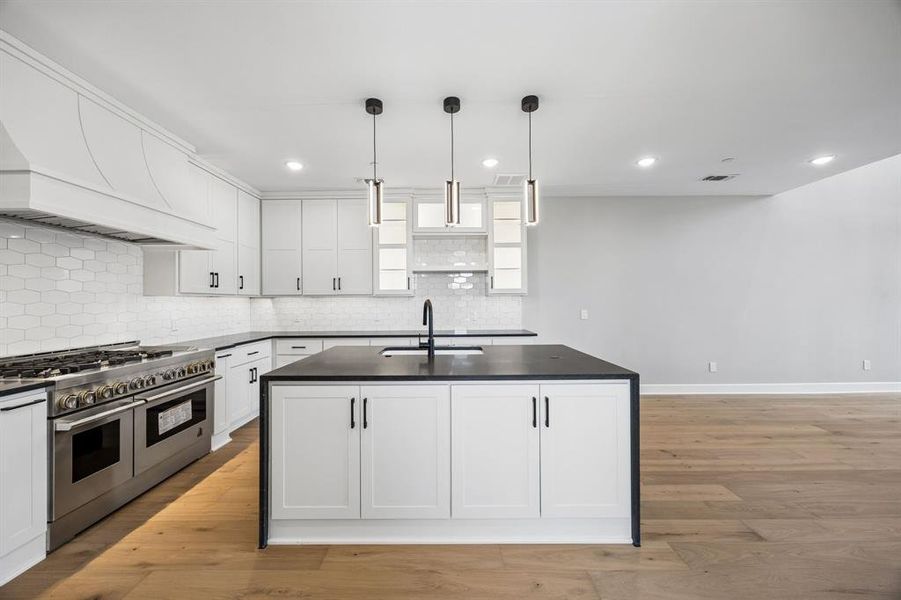 Kitchen with a center island with sink, range with two ovens, light wood finished floors, white cabinetry, and hanging light fixtures Kitchen with a center island with sink, range with two ovens, light wood finished floors, white cabinetry, and hanging light fixtures