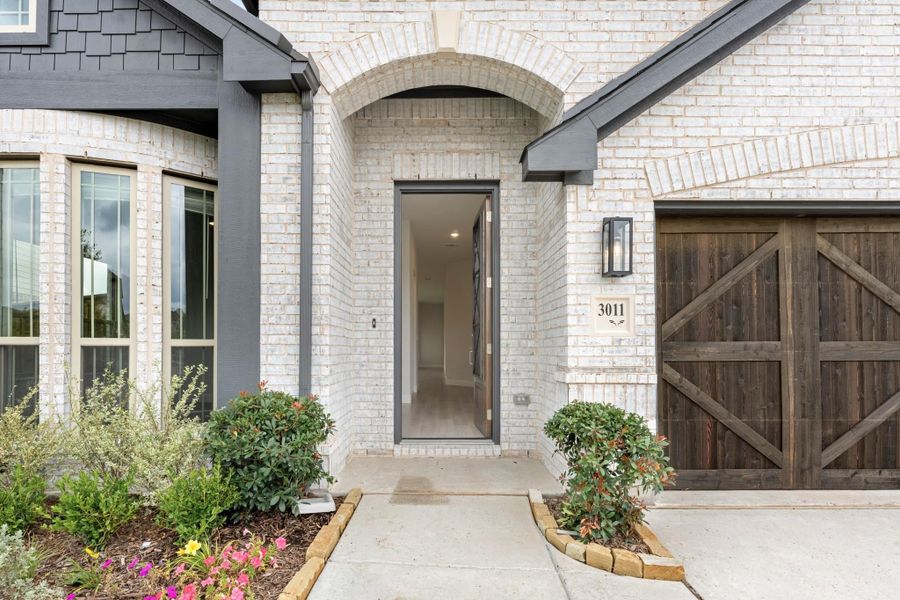Home entrance with brick arched doorway, open front door, wood garage door, and landscaped walkway with shrubs and flowers Home entrance with brick arched doorway, open front door, wood garage door, and landscaped walkway with shrubs and flowers