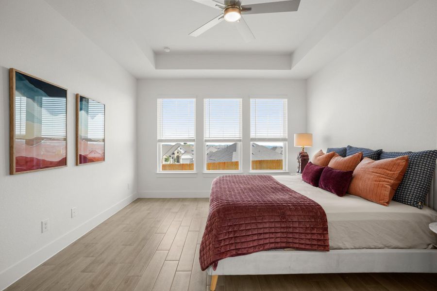 Natural light floods this primary bedroom featuring a tray ceiling, ceiling fan, and durable wood flooring.