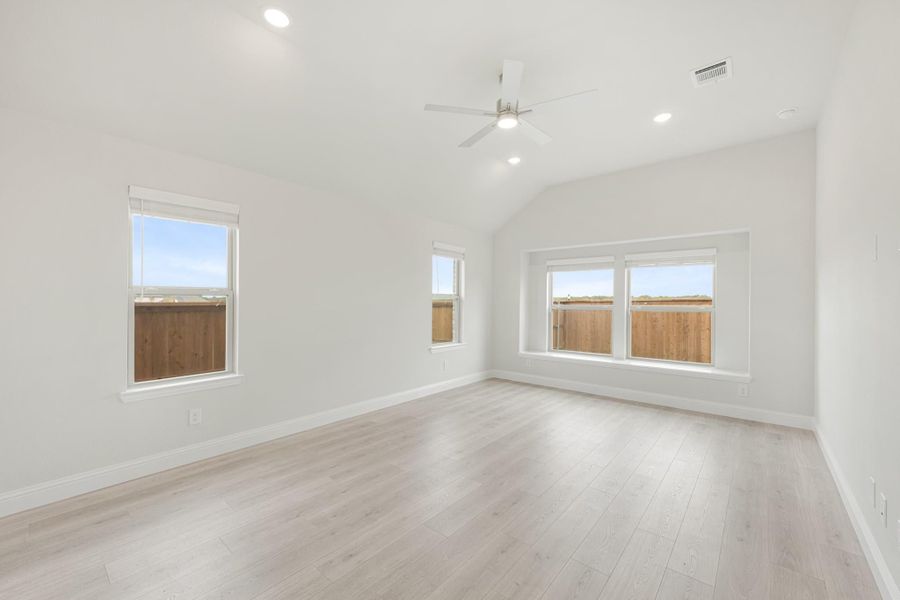 Empty bedroom with vaulted ceiling, ceiling fan, light wood floors, and multiple windows with natural light.