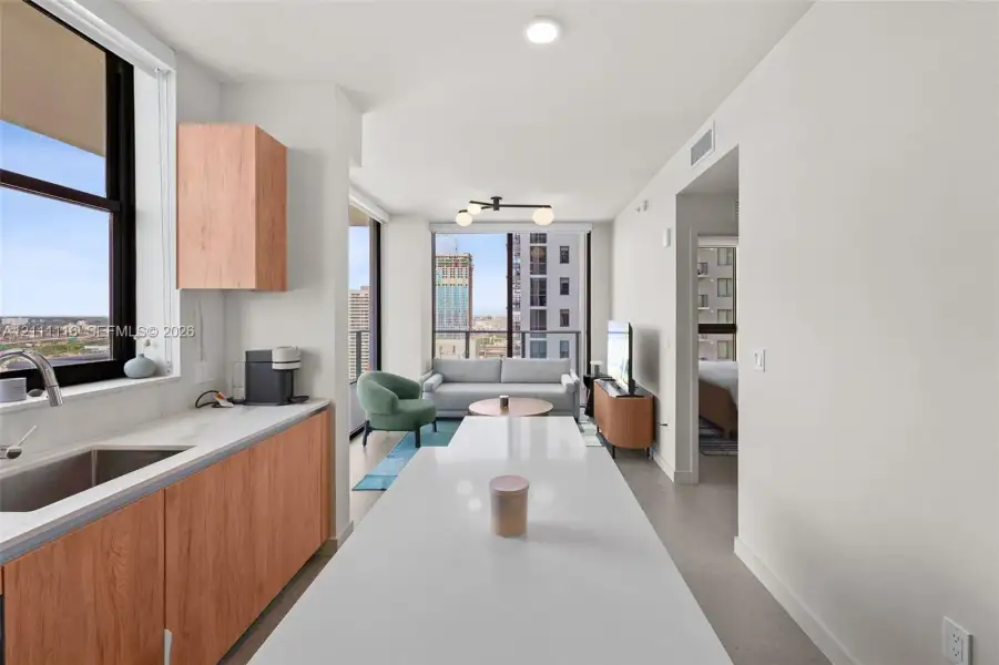Kitchen featuring wood-finish cabinetry, white solid-surface countertops, and an undermount stainless steel sink with a high-arch faucet, complemented by floor-to-ceiling windows and recessed lighting