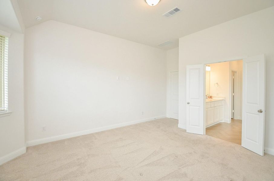 This bright, spacious primary bedroom features neutral carpeting, a high ceiling, and an en-suite bathroom with double doors, offering comfort and privacy.