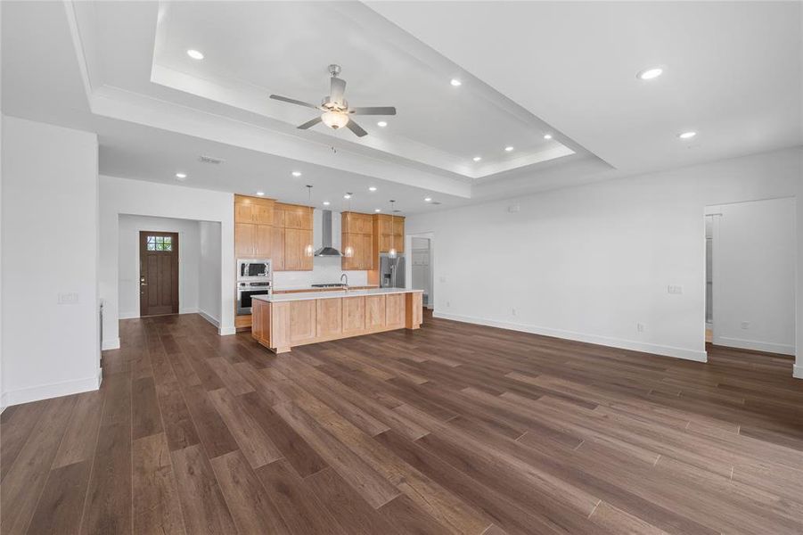 Open-concept living area featuring wood-finish flooring, a tray ceiling with recessed lighting and ceiling fan, and a kitchen with light wood cabinetry, stainless steel appliances, and a central island