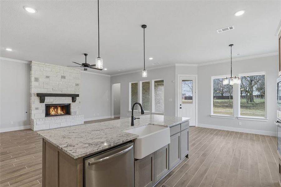 Kitchen featuring wood finish floors, stainless steel dishwasher, a stone fireplace, light stone counters, and hanging lights