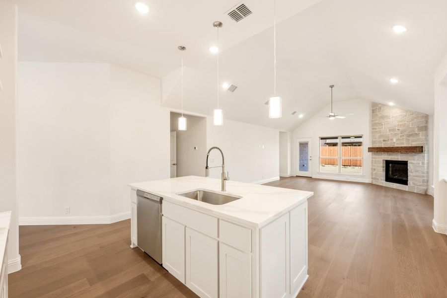 Open-concept kitchen with white island, quartz countertop, and pendant lights, opening to living room with stone fireplace Open-concept kitchen with white island, quartz countertop, and pendant lights, opening to living room with stone fireplace