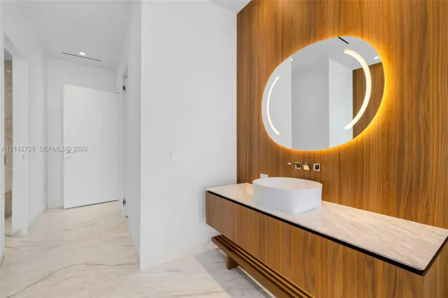 Floating wood-finish vanity with a stone countertop, white vessel sink, wall-mounted faucet, and custom wood-paneled wall featuring an integrated circular mirror