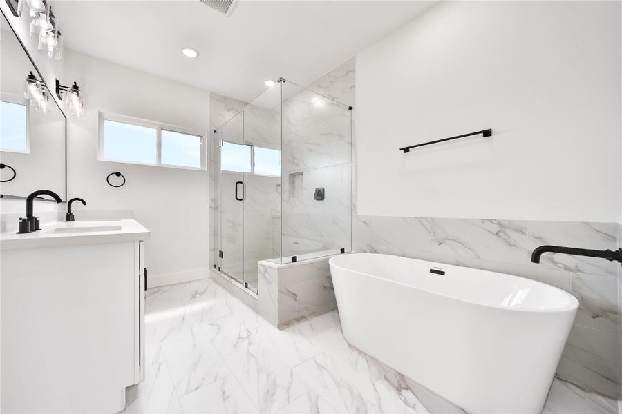 This bathroom features a modern, sleek design with a freestanding bathtub and a spacious glass-enclosed shower. It has elegant marble-style tiles, a double vanity with black fixtures, and ample natural light from a large window.