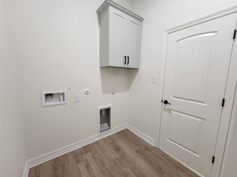 Laundry area featuring wood-finish flooring, white baseboards, a white shaker-style cabinet with black pull hardware, and an arched panel interior door with black hardware