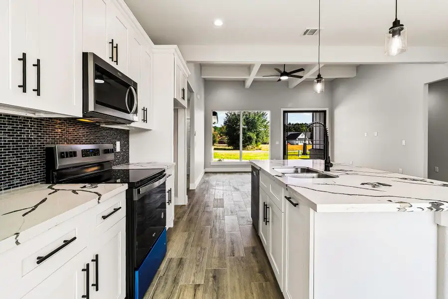 This modern kitchen features sleek white cabinetry, a stylish black backsplash, and elegant marble-patterned countertops. It includes stainless steel appliances, a central island with a sink, and pendant lighting. The open layout extends to a bright living area with large windows offering great natural light and views of the outdoors. This modern kitchen features sleek white cabinetry, a stylish black backsplash, and elegant marble-patterned countertops. It includes stainless steel appliances, a central island with a sink, and pendant lighting. The open layout extends to a bright living area with large windows offering great natural light and views of the outdoors.