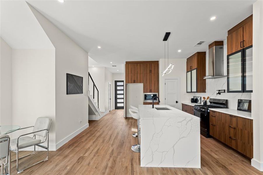 Kitchen featuring light stone counters, black appliances, wood finish cabinets, an island with sink, and hanging light fixtures