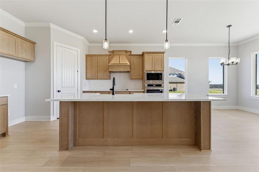Kitchen with ornamental molding, pendant lighting, light stone counters, custom range hood, and a center island with sink