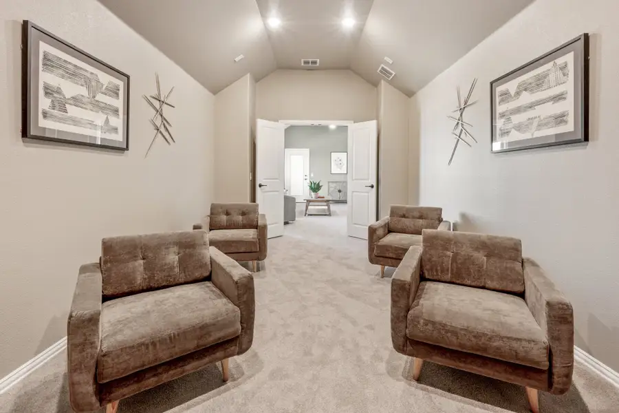 Sitting area with four brown armchairs, vaulted ceiling, carpet flooring, and framed wall art leading to an open doorway