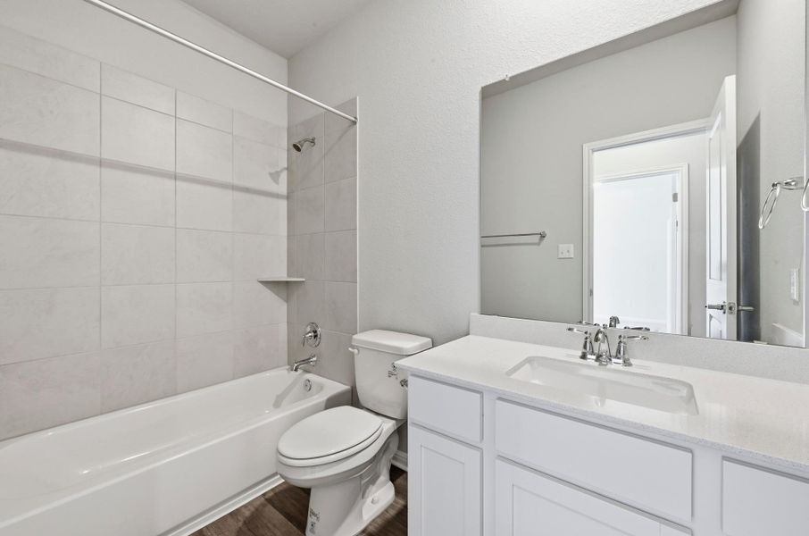 Contemporary bathroom featuring a white vanity with integrated sink, a large wall-mounted mirror, a white bathtub with subway tile surround, a toilet, and wood-finish flooring Contemporary bathroom featuring a white vanity with integrated sink, a large wall-mounted mirror, a white bathtub with subway tile surround, a toilet, and wood-finish flooring