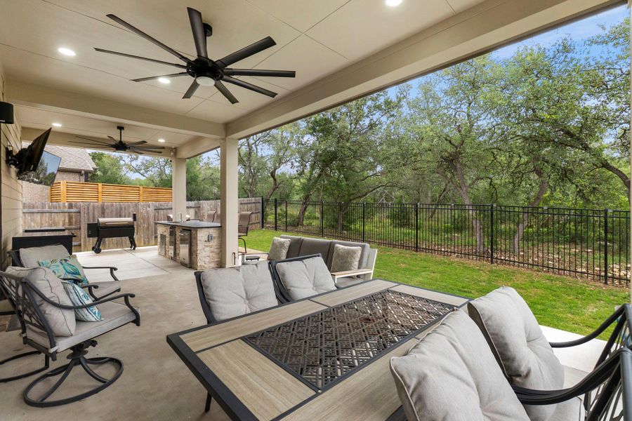Covered patio featuring a built-in outdoor kitchen with stone veneer and a counter surface, two ceiling fans, recessed lighting, and a metal fence bordering a natural treeline