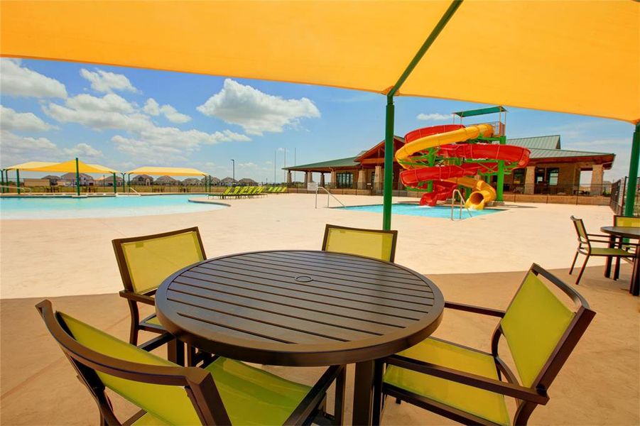 Shaded outdoor patio area featuring multiple tables and chairs, overlooking a community pool with a shallow entry, a water slide structure, and a clubhouse building