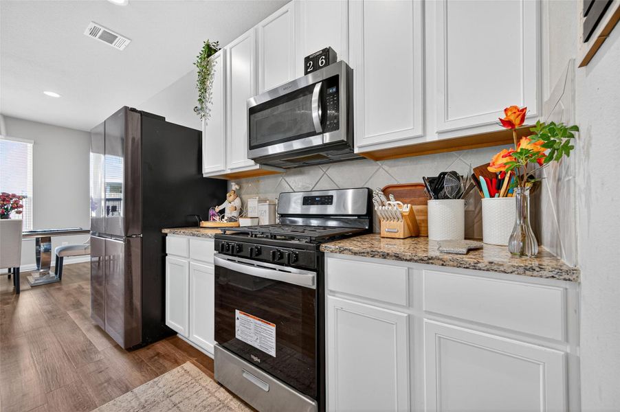 This kitchen features sleek white cabinetry, granite countertops, and stainless steel appliances, including a microwave and a gas stove. It offers a modern, clean look with ample natural light and a view into a dining area with wood flooring.