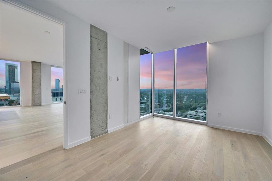 Step into this sleek and contemporary bedroom featuring floor-to-ceiling windows that frame spectacular views of the city and beyond. The natural light floods the room, enhancing the airy ambiance created by the light wood flooring and neutral color palette. The exposed concrete accent wall adds an industrial touch to the minimalist design. *Linden Residence No. 2105 Shown Here