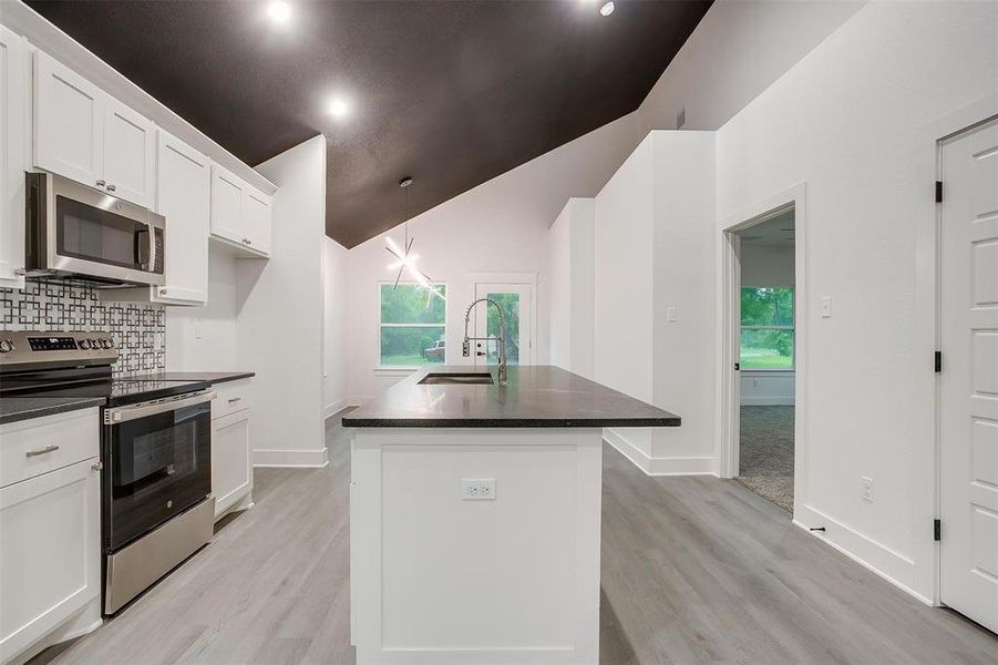 Kitchen featuring stainless steel appliances, dark countertops, an island with sink, white cabinetry, and high vaulted ceiling